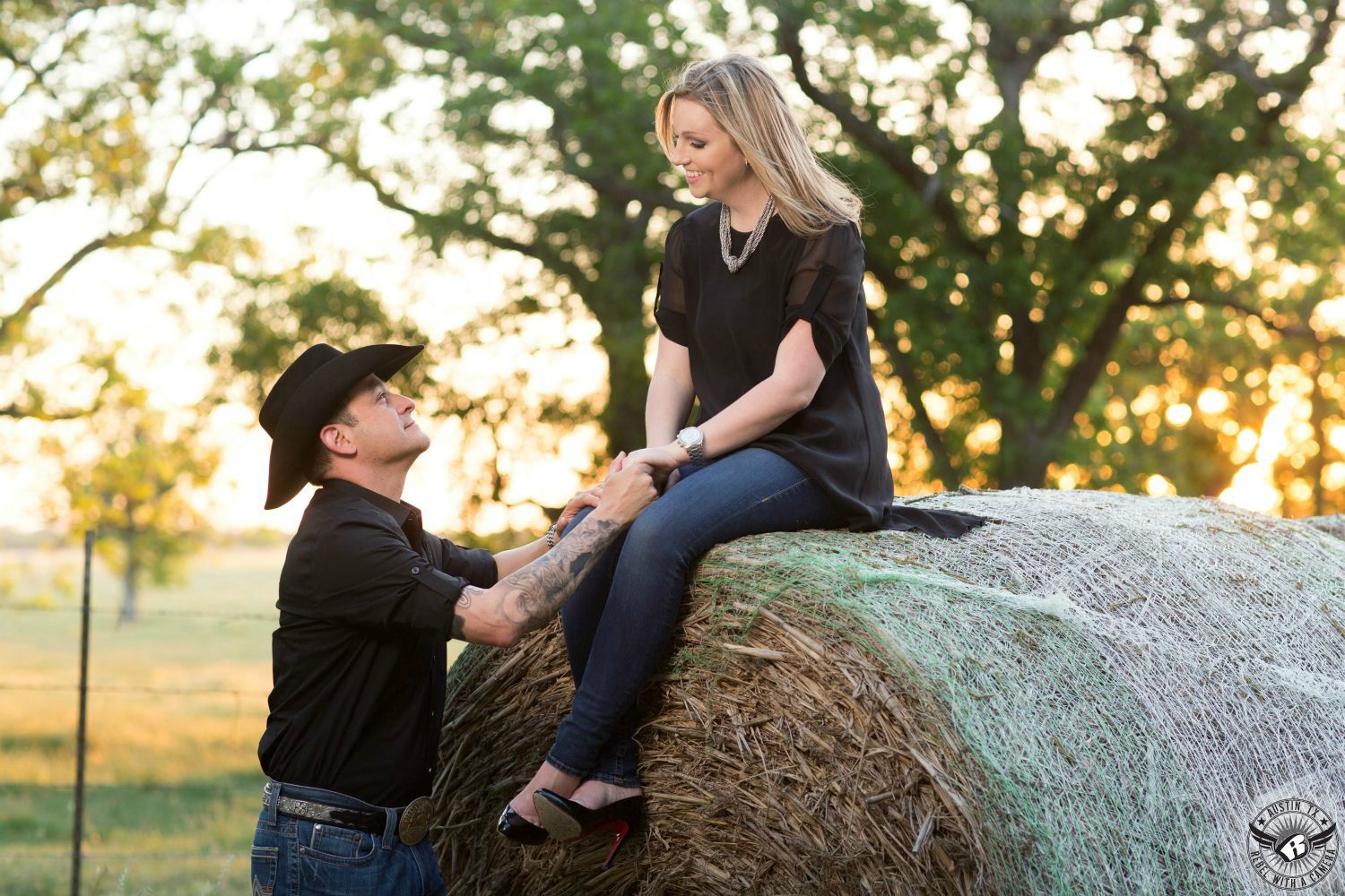Blond lady in a flowing black blouse with short sleeves, blue jeans and black heals sits on a large round hay bail  holding the hand of a tattooed Latino guy with a black cowboy hat wearing a black dress shirt with rolled up sleeves and blue jeans with a large belt buckle while looking up at the bride with a large warm glow in the background from the sunset through trees in this dreamy engagement portrait in Georgetown Texas in rural Austin. 